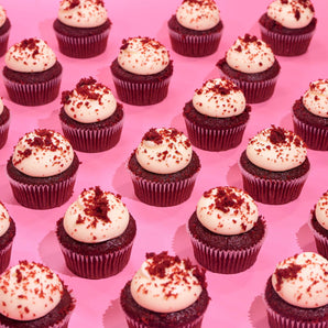 Row of Crumbs & Doilies Red Velvet Cupcakes with cream cheese frosting and red velvet crumbs on pink background